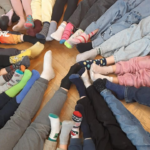 Zbliżenie na nogi i stopy dzieci ułożone w kręgu na drewnianej podłodze. Każde dziecko ma inne, kolorowe skarpetki.