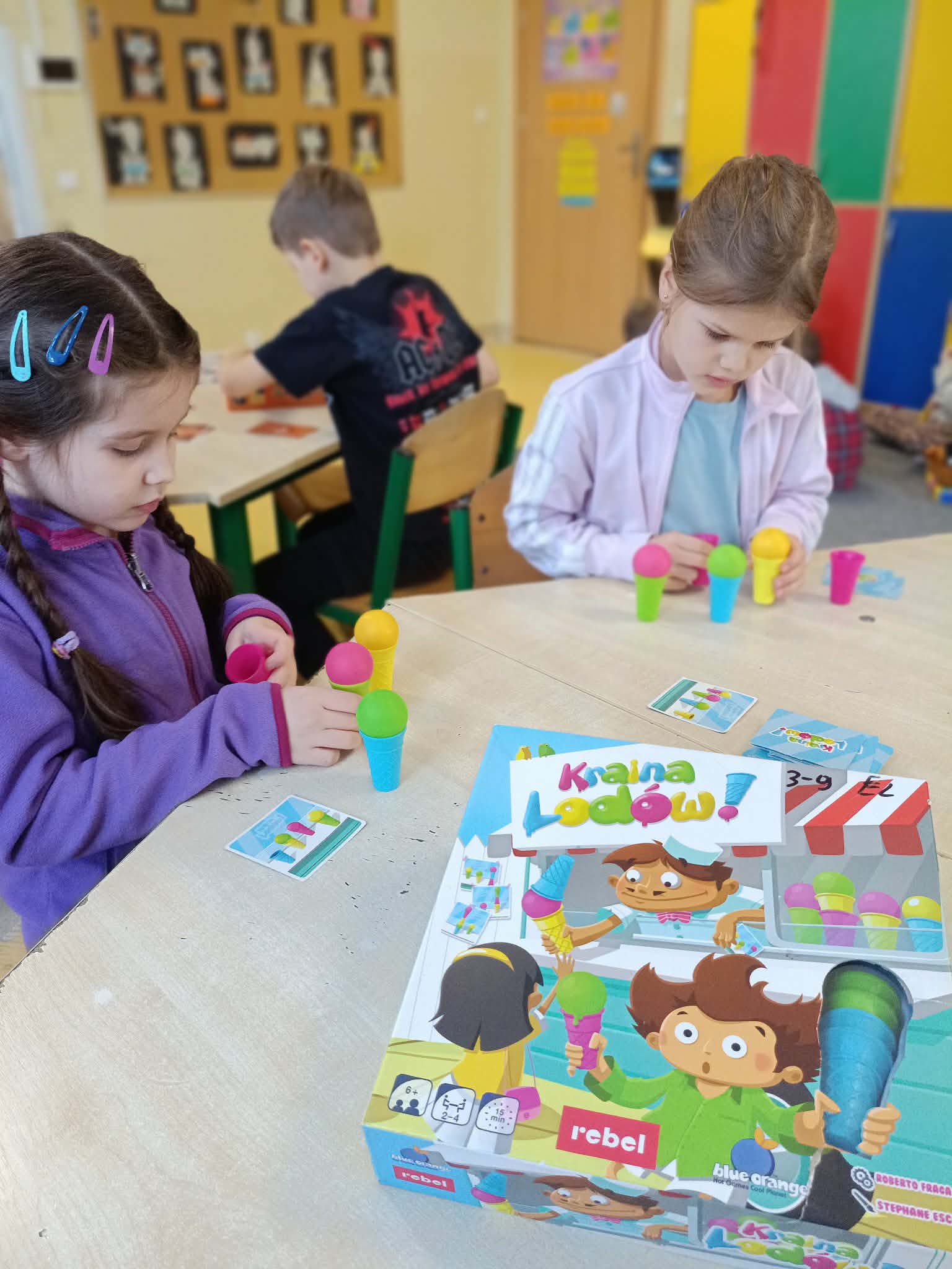 Dwie dziewczynki siedzą przy stoliku w sali i układają kolorowe kulki lodów na plastikowych rożkach według kart zadań w grze planszowej „Kraina Lodów”. Na stole leży pudełko z grą.