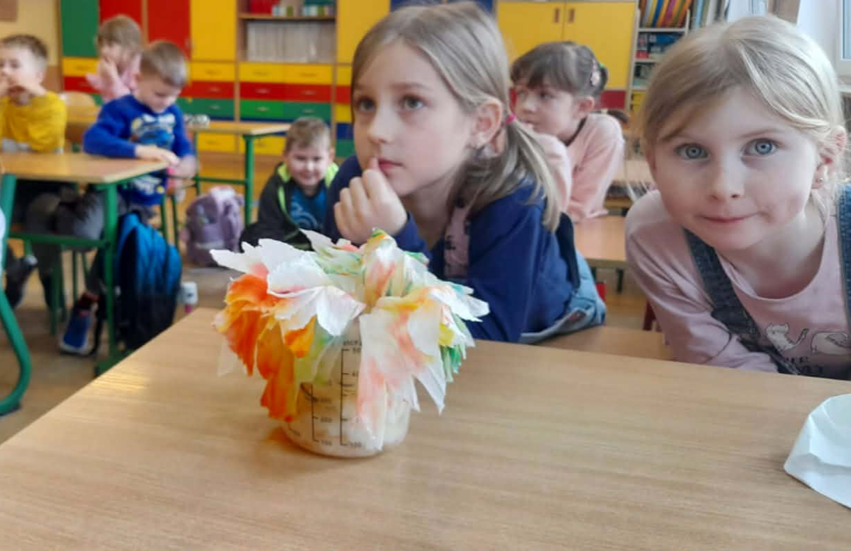 Grupa dzieci siedzi w klasie przy ławkach. Na pierwszym planie stoi słoik z kolorowymi, barwionymi papierowymi kwiatami. Dzieci w tle patrzą w różne strony – część jest skupiona, inne rozmawiają. W tle kolorowe szafki.