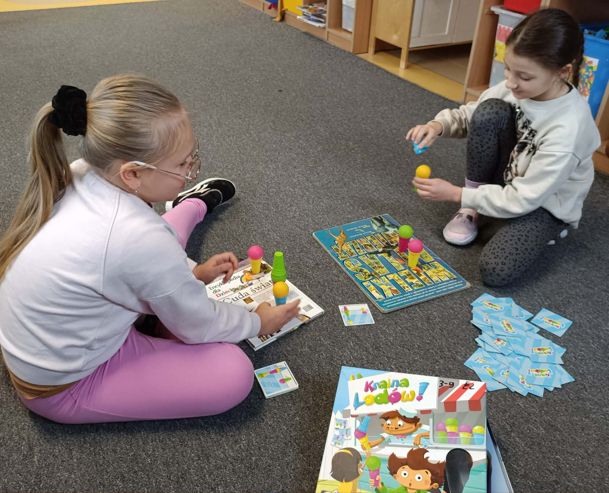Dwie dziewczynki siedzą na dywanie i grają w grę „Kraina Lodów”, układając kolorowe kulki lodów na rożkach zgodnie z kartami zadań. Obok leży pudełko z grą i stos kart.