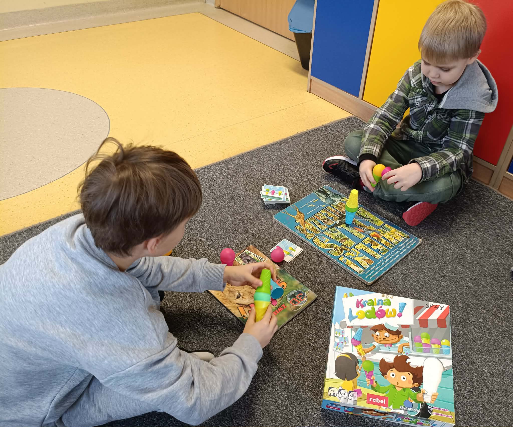 Dwóch chłopców siedzi na dywanie i układa elementy gry „Kraina Lodów”, dopasowując kolorowe kulki lodów do rożków według kart zadań. Obok znajduje się pudełko z grą.