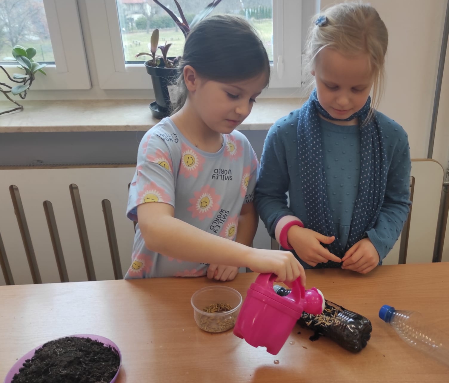  Dwie dziewczynki stoją za stołem, na którym leży pusta butelka, talerzyk z ziemią i nasiona w pudełku. Jedna z dziewczynek trzyma w dłoni konewkę. W tle okno, parapet i kwiaty doniczkowe.