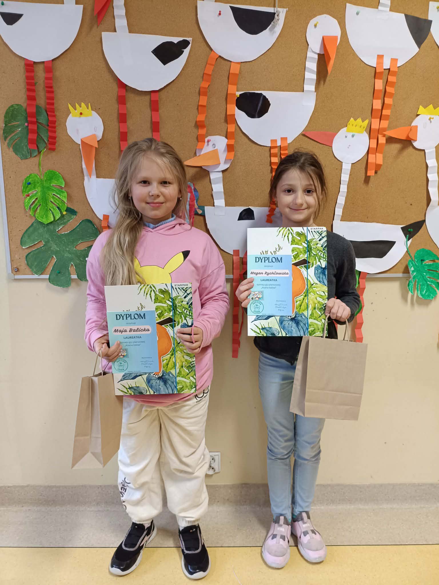 Dwie dziewczynki stoją przed tablicą z papierowymi pracami plastycznymi przedstawiającymi bociany. Trzymają dyplomy oraz torby z nagrodami.
