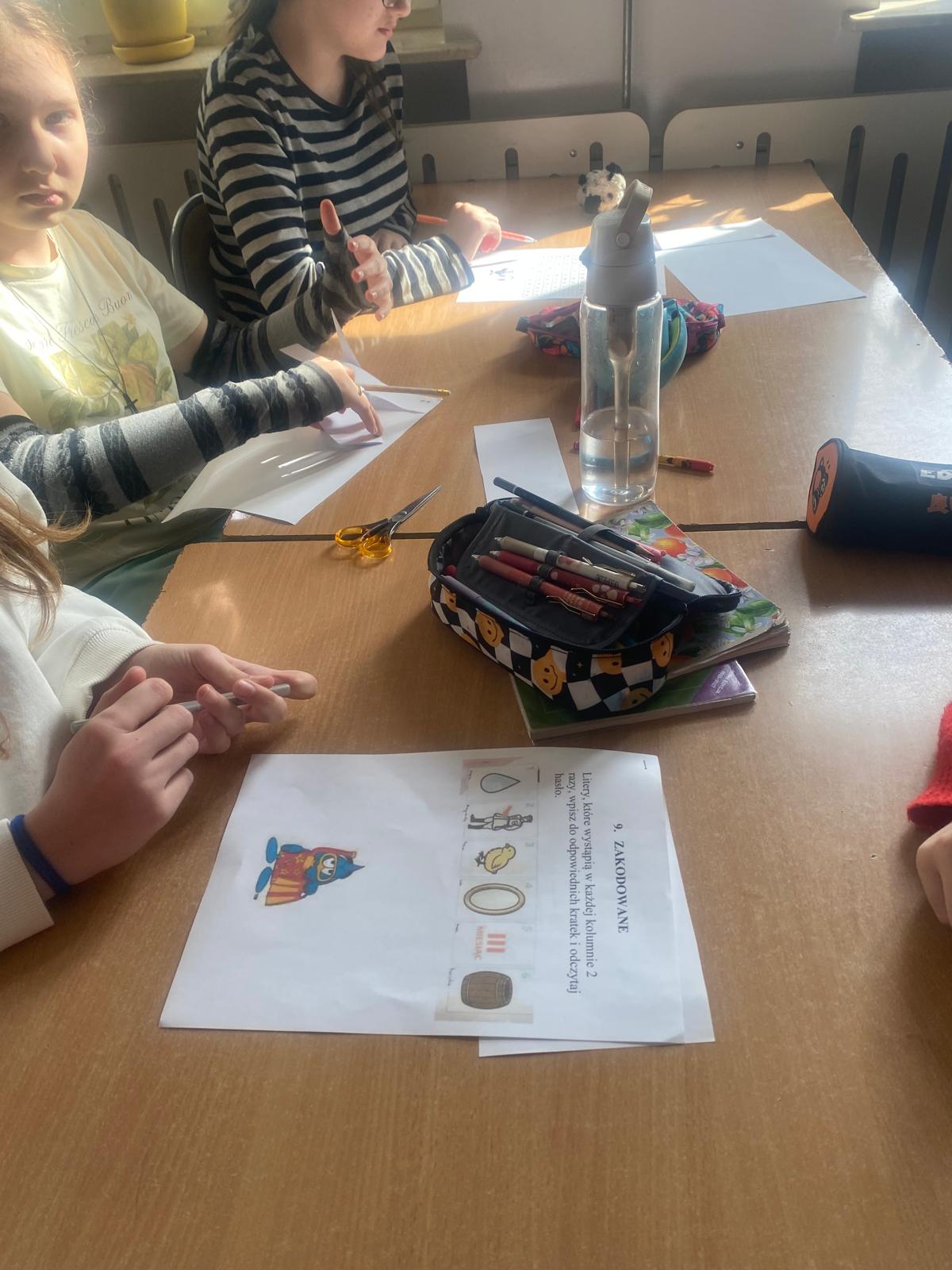 Dzieci siedzą przy stole w klasie i wykonują zadanie plastyczne z kartkami, kredkami i nożyczkami.