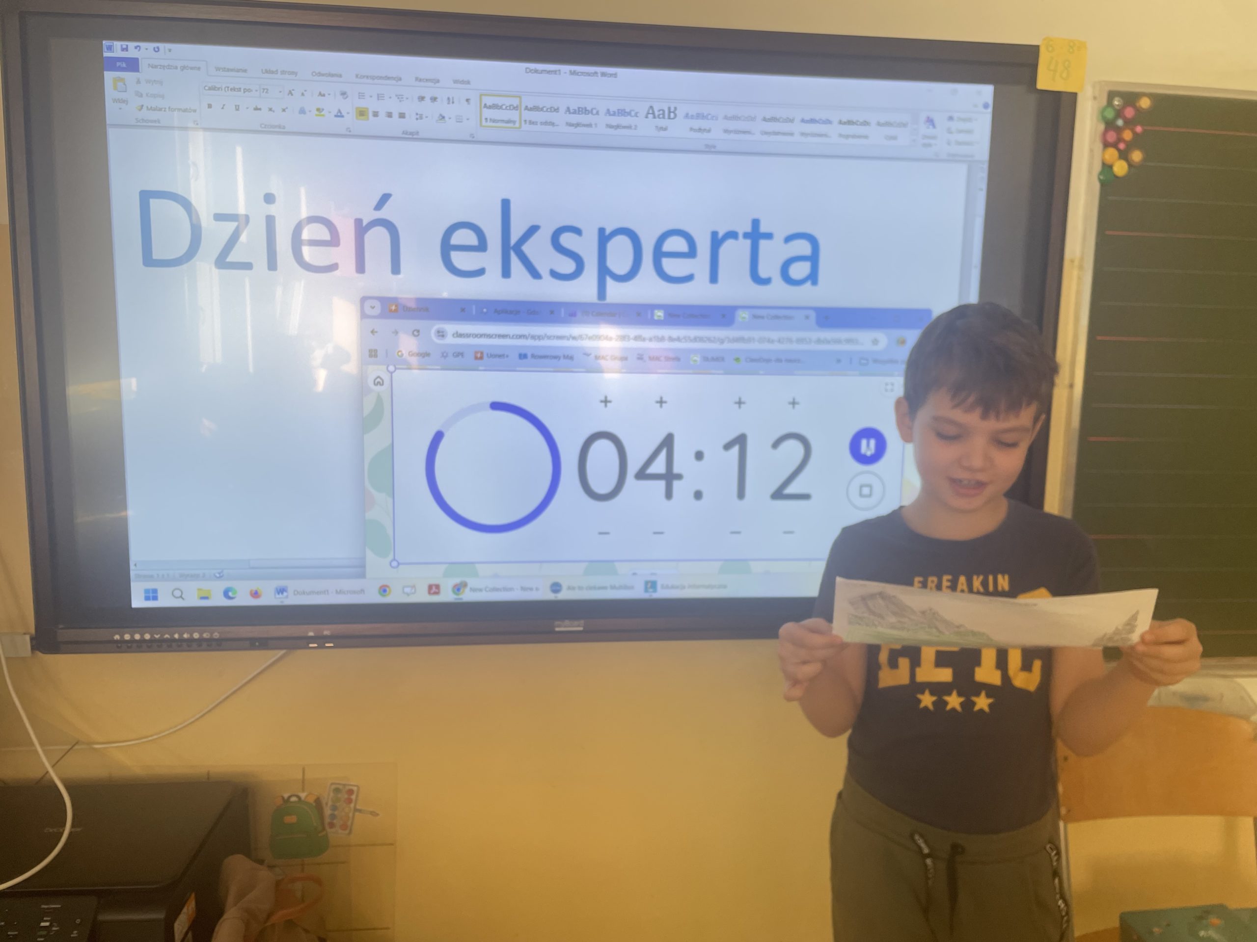 Uczeń stoi przed ekranem multimedialnym, w rękach trzyma swoją pracę plastyczną. 