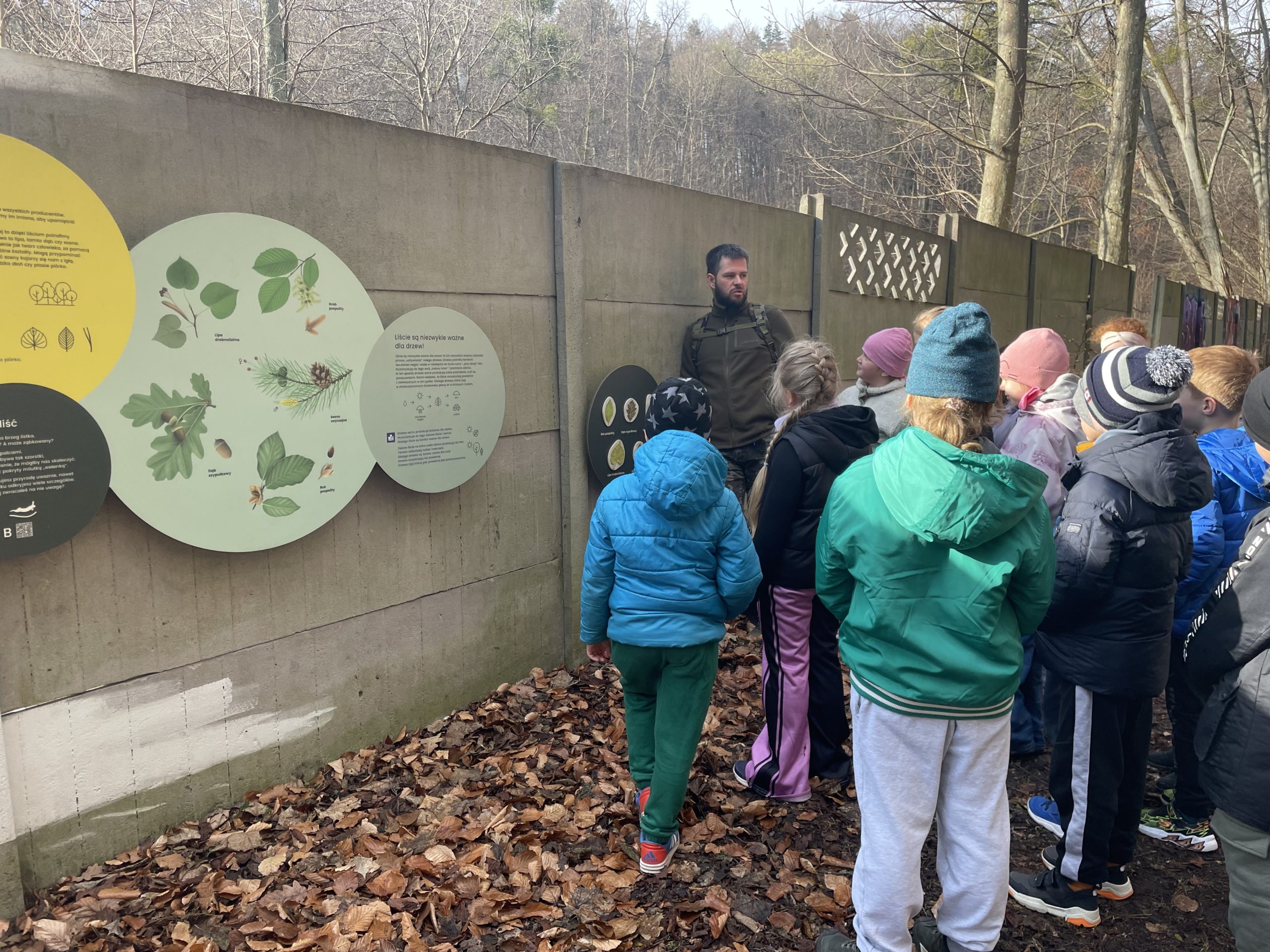Edukator i grupa uczniów stoi przy informacji o gatunkach roślin.