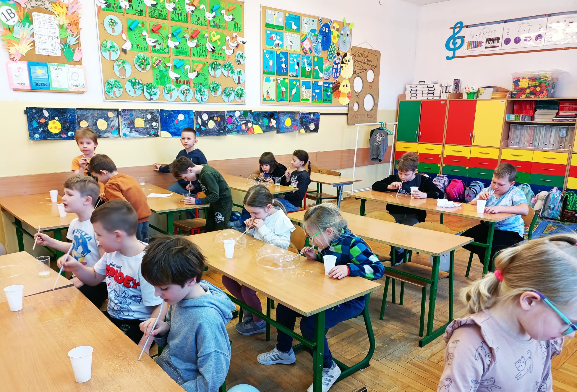 Dzieci siedzą przy ławkach i wykonują doświadczenie z tworzeniem baniek mydlanych przy użyciu słomek i kubków.