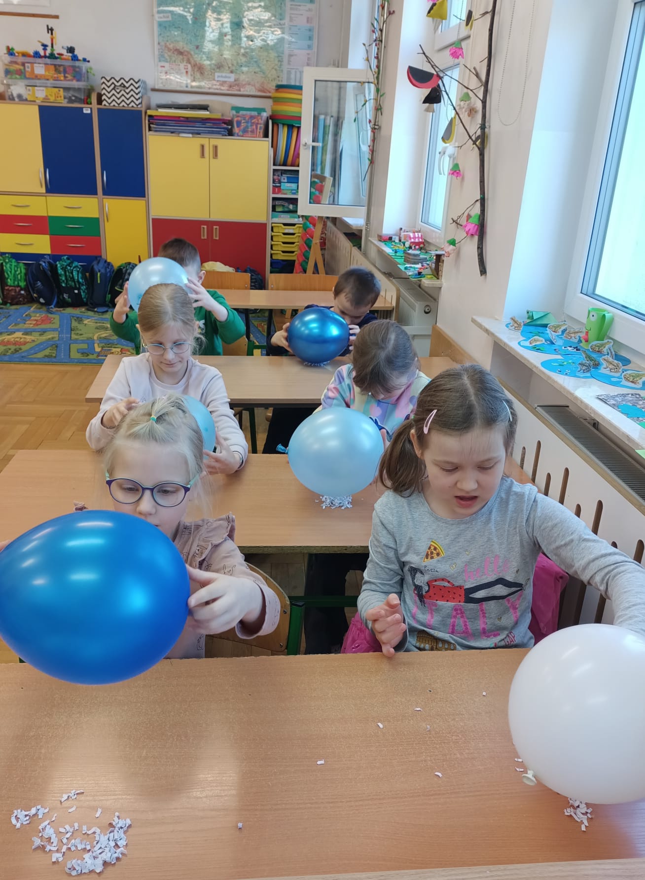 Uczniowie siedzą przy ławkach z balonami, skupieni na doświadczeniu polegającym na przyciąganiu skrawków papieru.