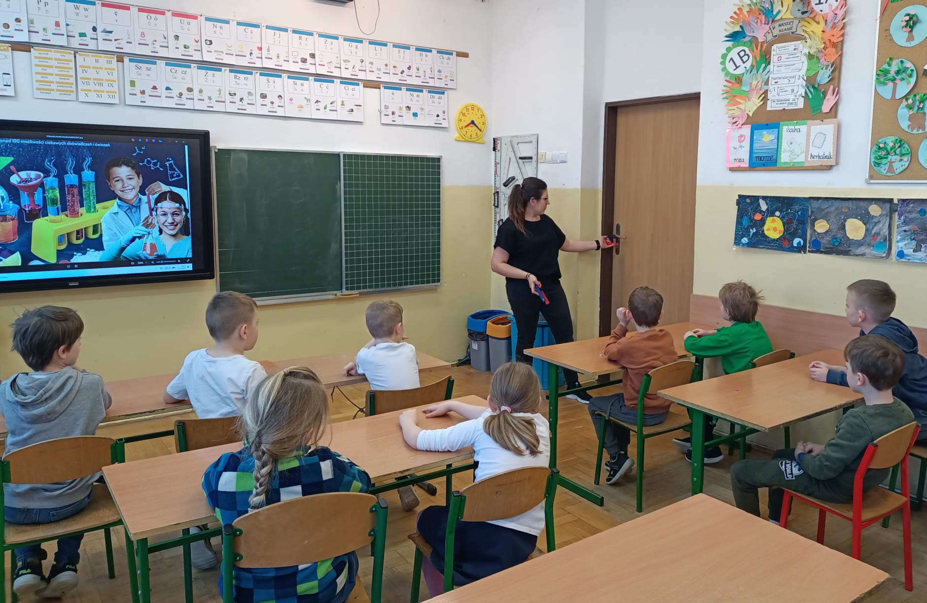 Nauczycielka prowadzi zajęcia w klasie, a uczniowie siedzą w ławkach i oglądają prezentację dotyczącą eksperymentów.
