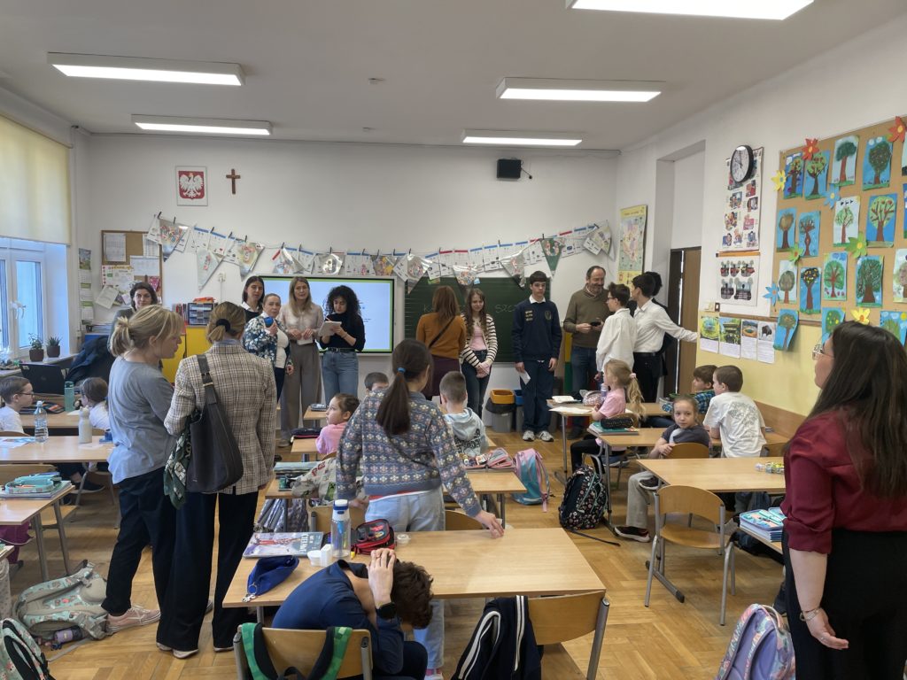 W klasie dzieci siedzą w ławkach a wokół nich są osoby które oglądaja co robią dzieci. Widać dużo kolorowych dekoracji