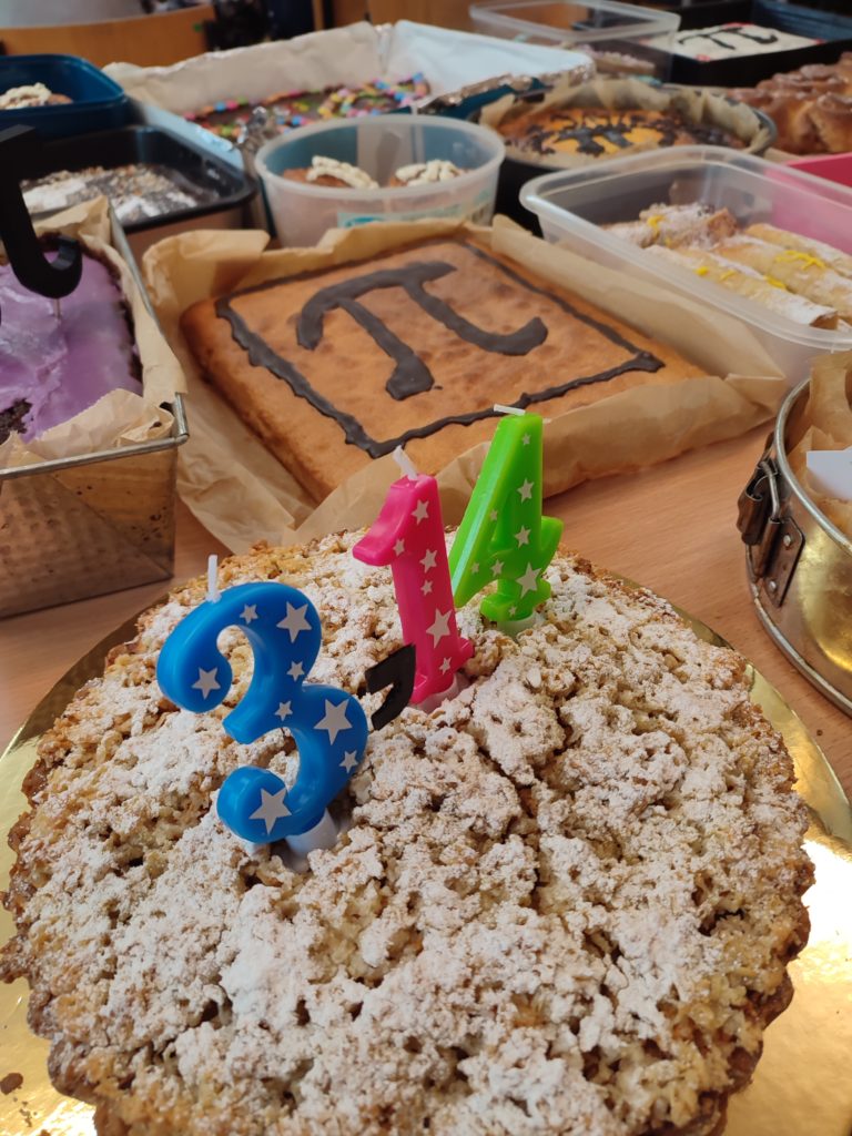 Zbliżenie na okrągłe ciasto posypane cukrem pudrem z zapalonymi świeczkami w kształcie cyfr 3, 1 i 4, w tle inne wypieki z symbolem π.