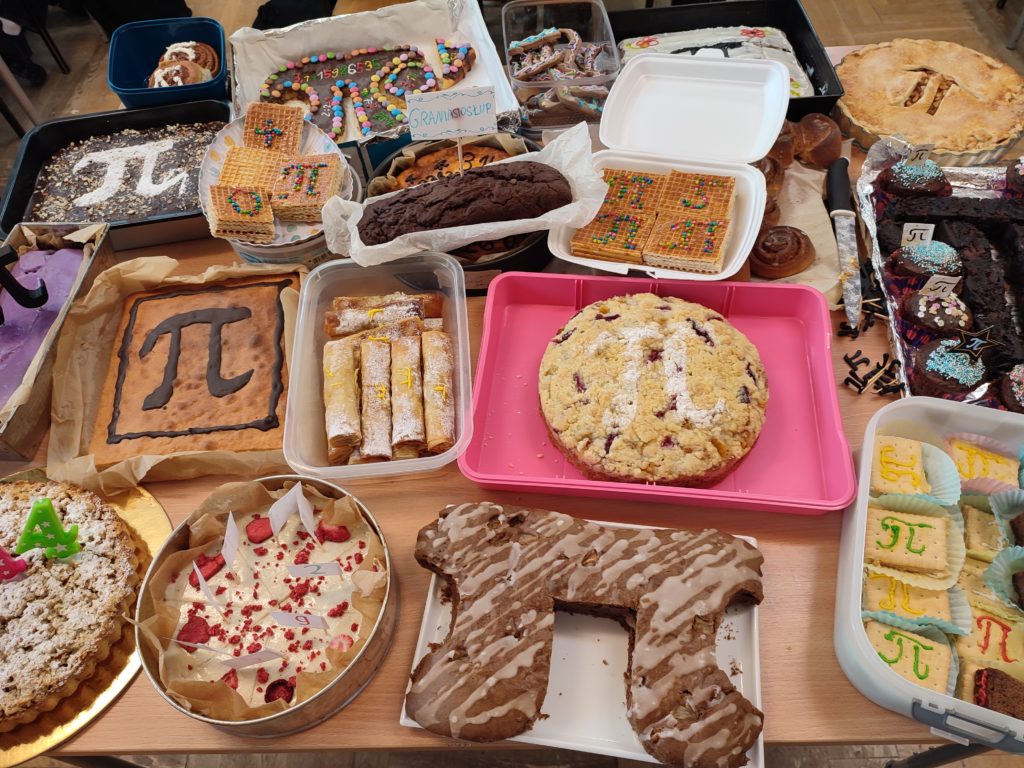 Różnorodne ciasta i ciasteczka ułożone na stole, w tym duże ciasto w kształcie symbolu π oraz wypieki ozdobione kolorowymi posypkami i cyframi.
