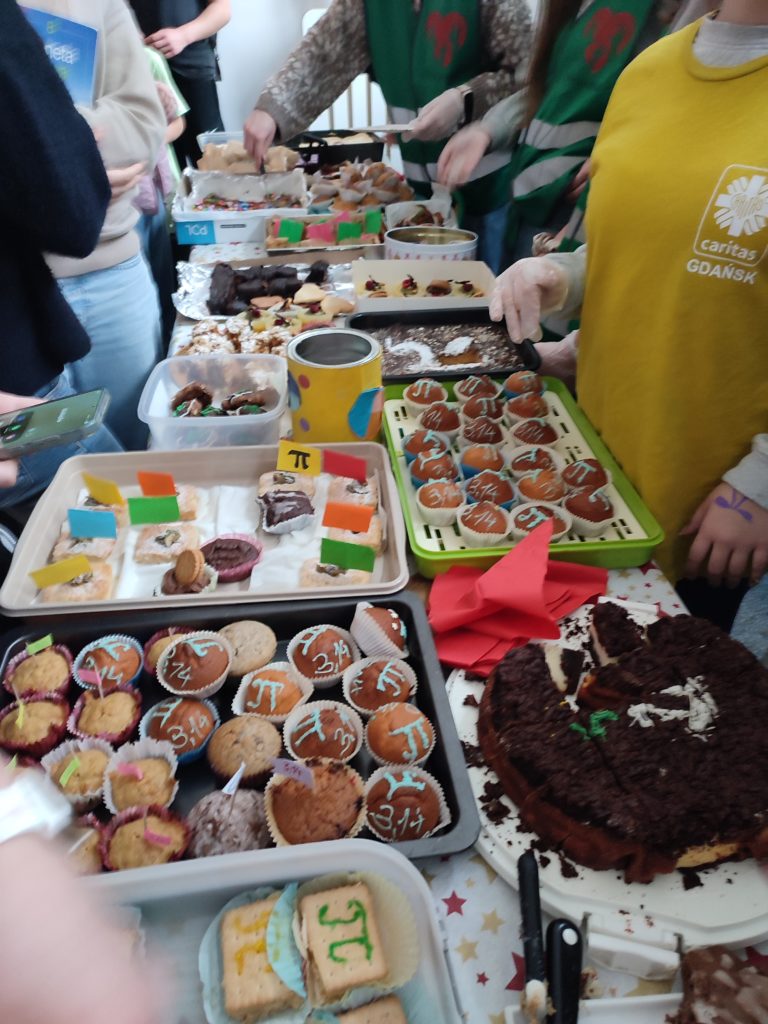 Zbliżenie na stoisko z wypiekami – muffiny, ciasta i ciasteczka z dekoracjami przedstawiającymi liczbę π, podawane przez uczniów w rękawiczkach.