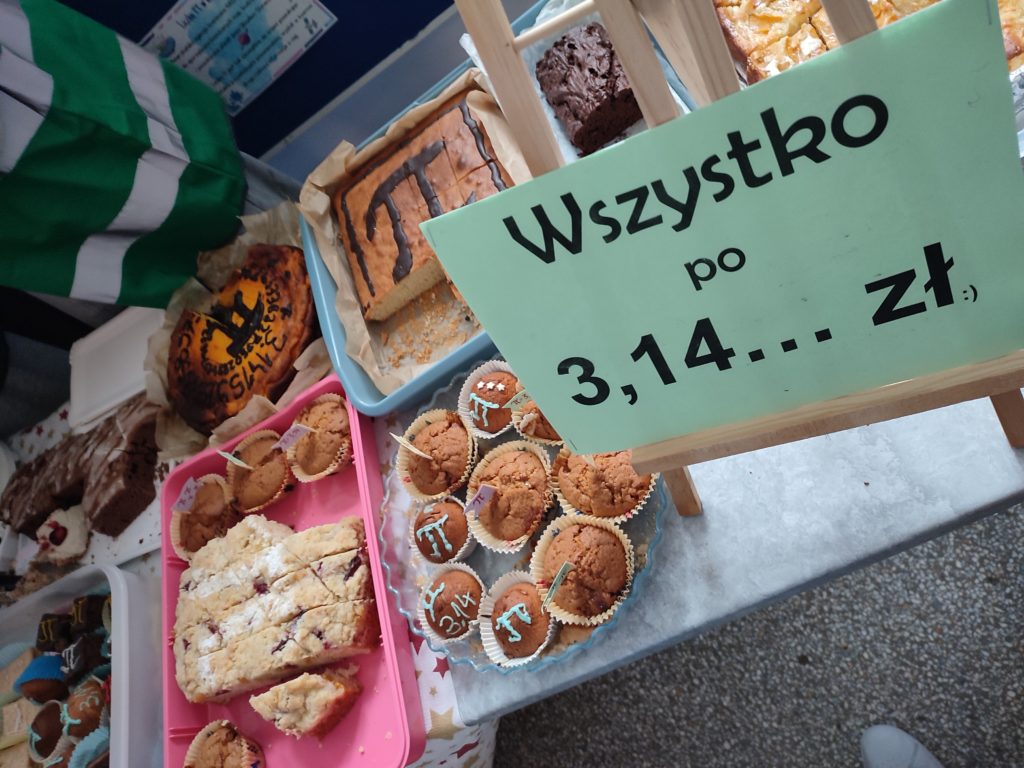 Stoisko z wypiekami i tabliczką z napisem „Wszystko po 3,14 zł”, obok różne ciasta i muffiny ozdobione symbolem π.
