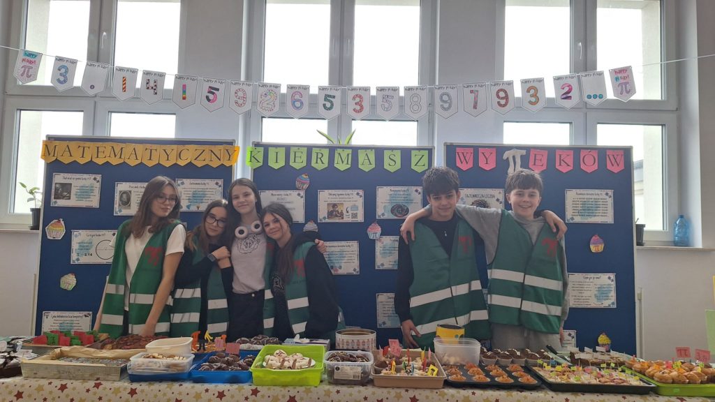 Grupa uczniów stojących za stołem z wypiekami podczas matematycznego kiermaszu, nad nimi girlanda z cyframi rozwinięcia liczby π i plansze informacyjne.
