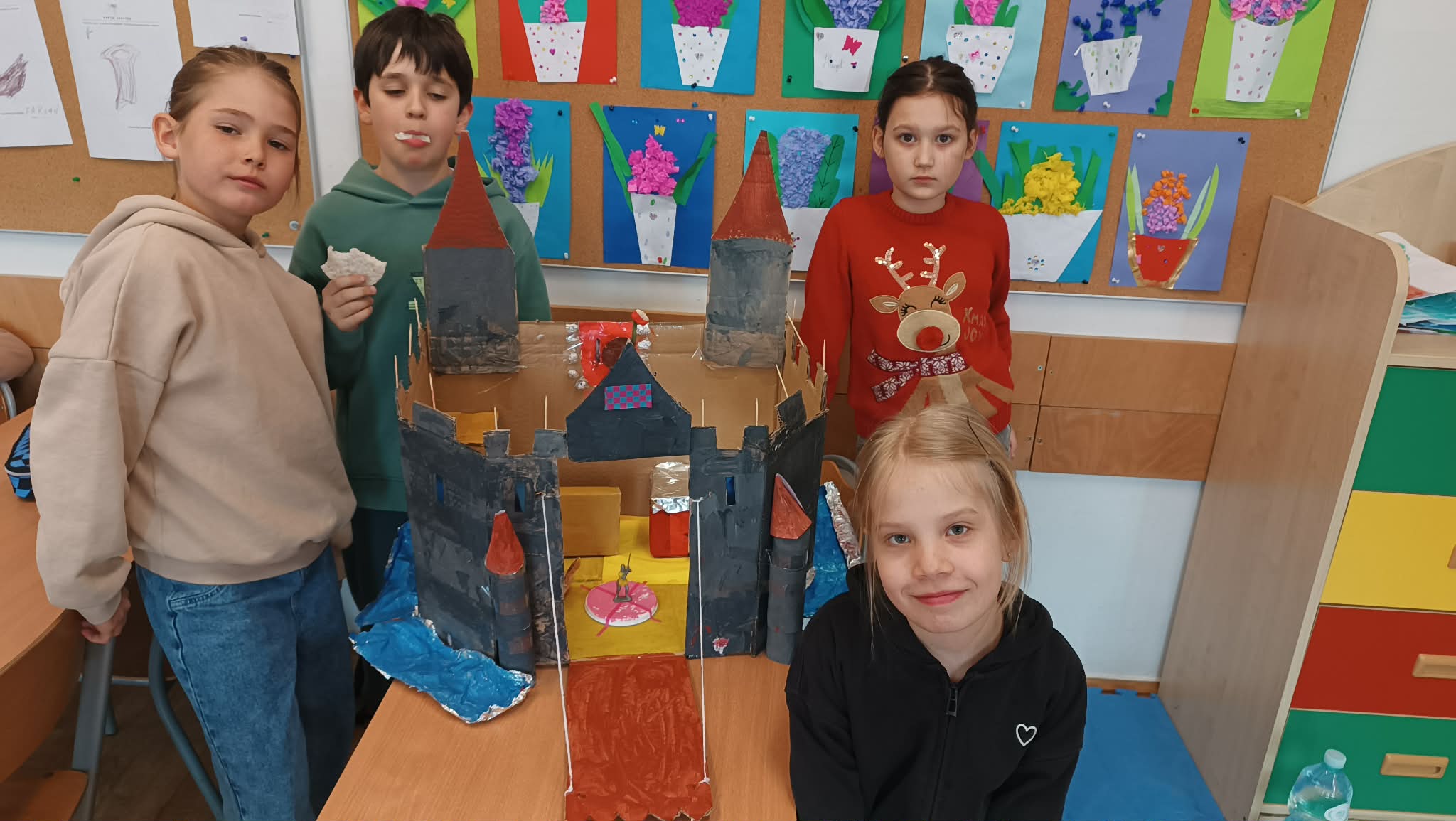 Dzieci stoją przy makiecie zamku z wieżami i mostem zwodzonym, którą samodzielnie przygotowały na zajęciach plastycznych. Praca wyróżnia się starannością wykonania oraz pomysłowym wykorzystaniem różnych materiałów.