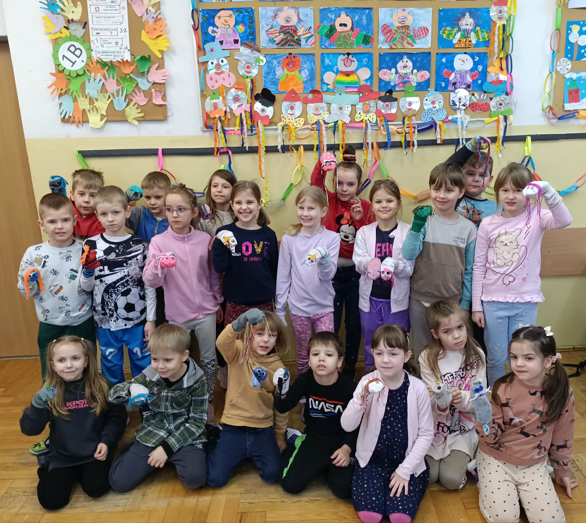  Grupa dzieci stoi /siedzi w sali lekcyjnej, trzymając wykonane przez siebie pacynki ze skarpetek. W tle znajdują się tablice z pracami plastycznymi i kolorowymi dekoracjami.