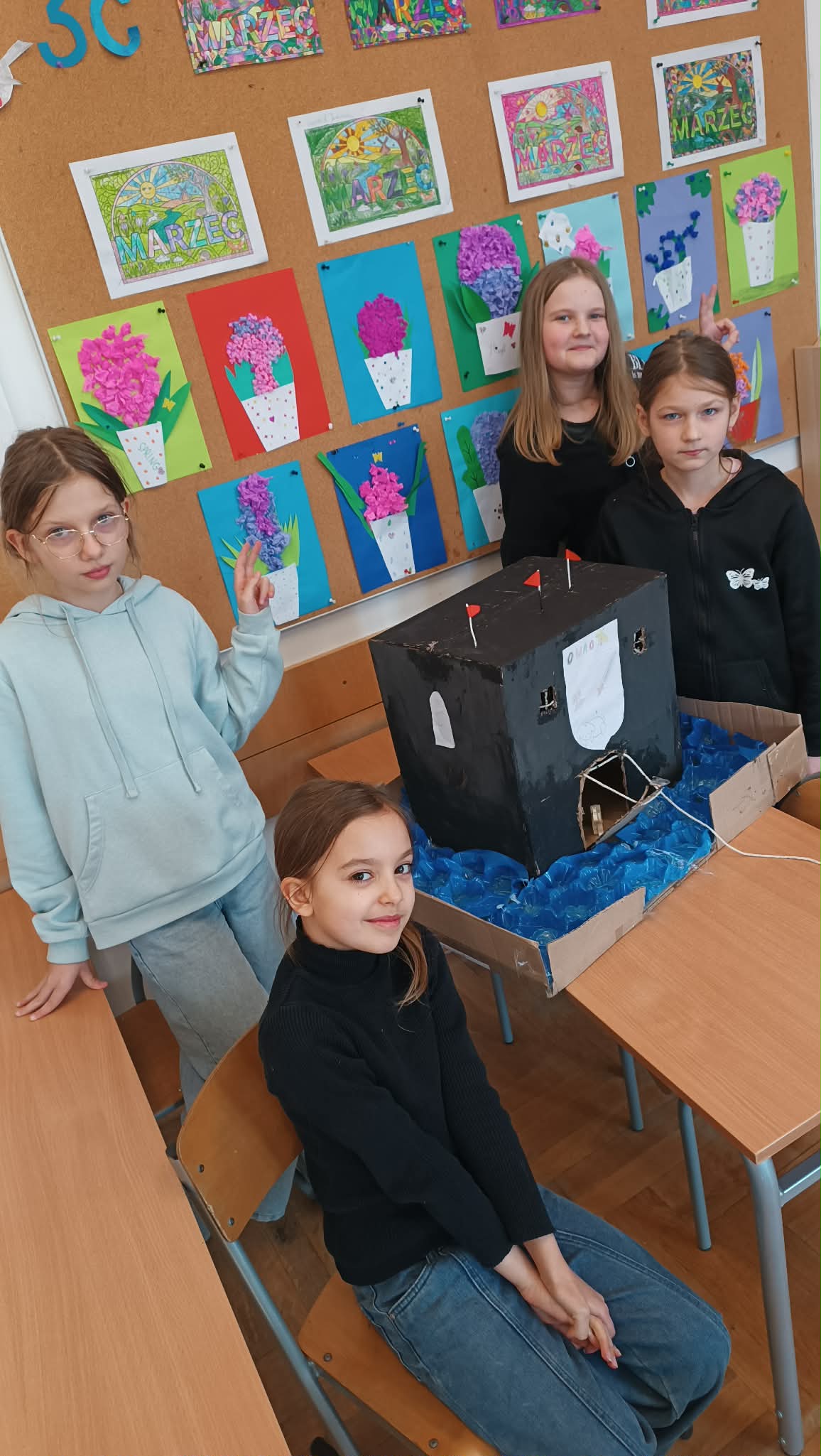 Cztery uczennice pozują w klasie przy własnoręcznie wykonanej makiecie czarnego zamku z mostem zwodzonym. W tle widać tablicę korkową z kolorowymi pracami plastycznymi z napisem „MARZEC”.