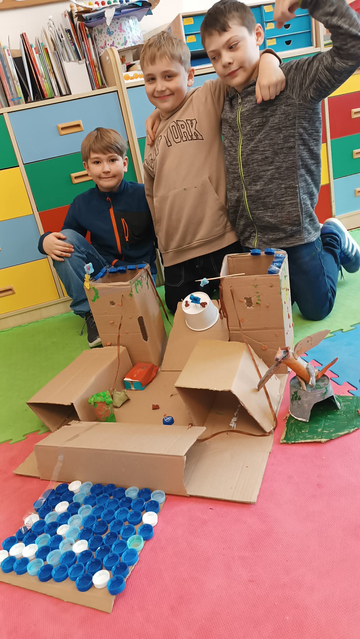 Grupa chłopców prezentuje makietę zamku wykonaną z kartonów i materiałów plastycznych podczas zajęć szkolnych. Projekt zawiera elementy takie jak wieże, dziedziniec oraz dekoracje wykonane z recyklingowych materiałów.