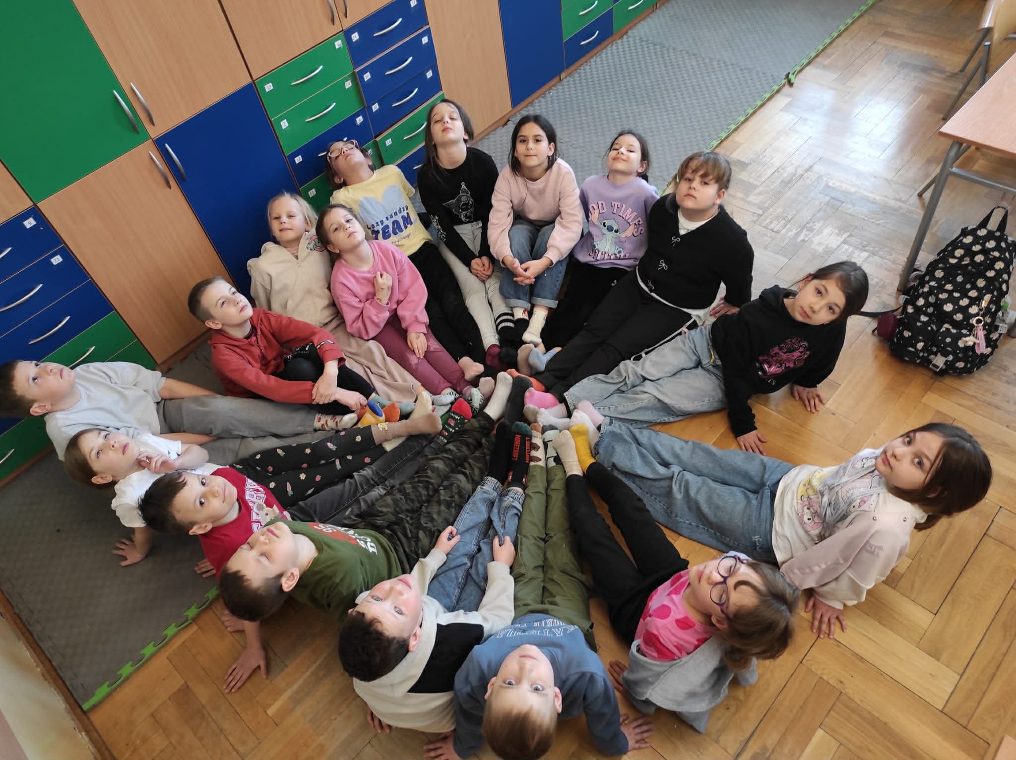 Grupa dzieci siedzi w kręgu w sali lekcyjnej. Dzieci siedzą z wyciągniętymi nogami do środka, tworząc okrąg. 