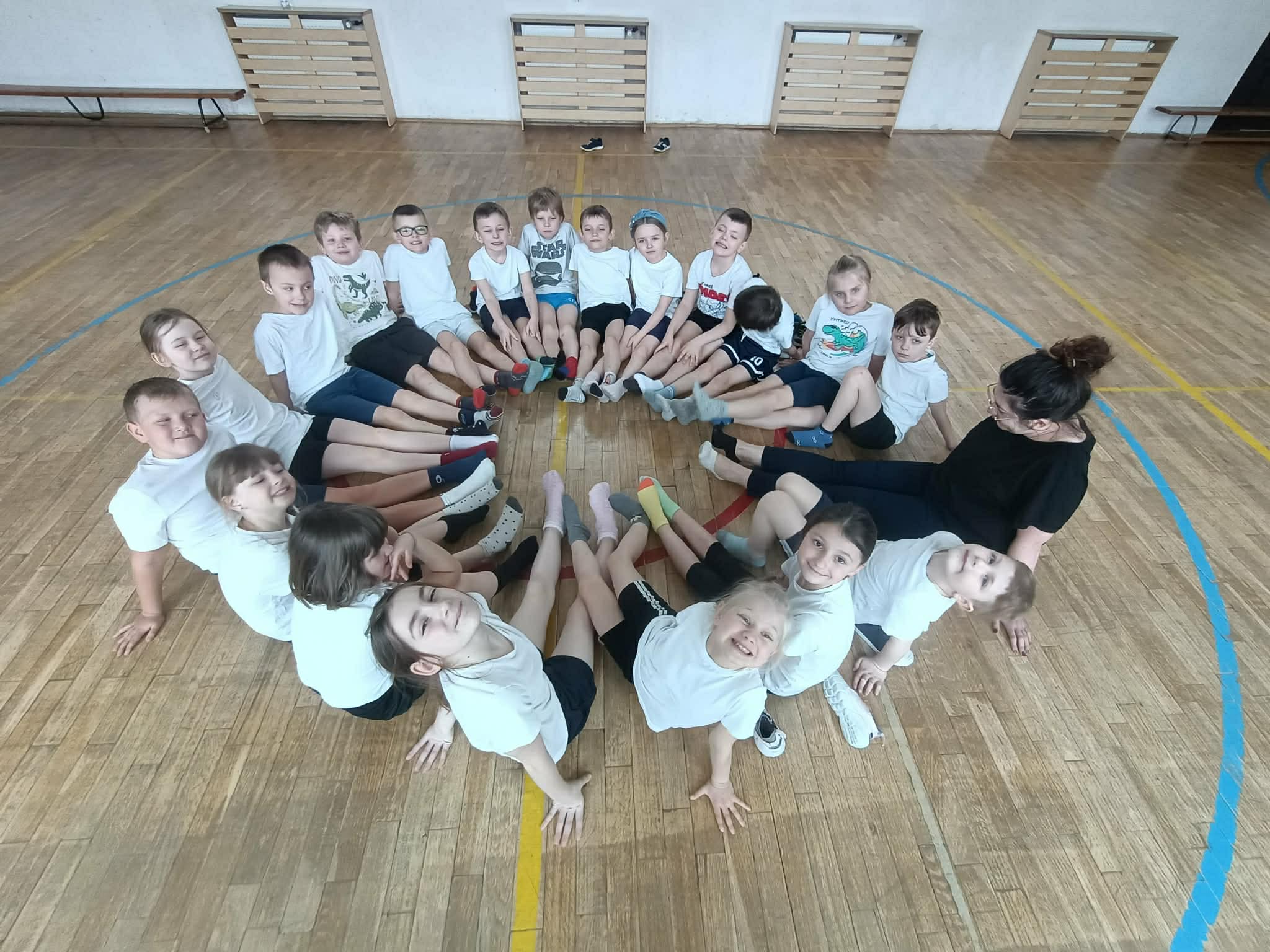 Grupa dzieci siedzi w kręgu na sali gimnastycznej razem z nauczycielką. Dzieci mają na sobie głównie białe koszulki i siedzą z wyciągniętymi nogami do środka, tworząc okrąg. 