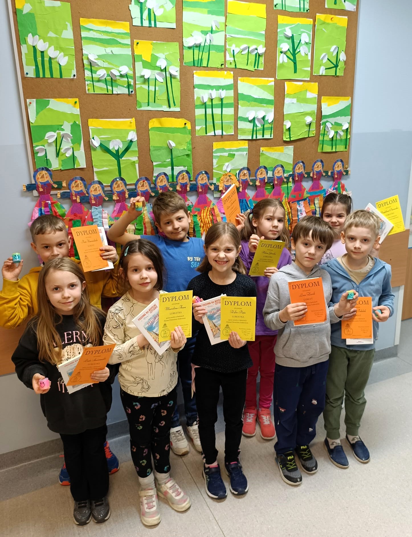 Grupa dzieci stoi na korytarzu przed tablicą z pracami plastycznymi przedstawiającymi wiosenne kwiaty. Każde dziecko trzyma kolorowy dyplom i książkę. Dzieci stoją w dwóch rzędach i uśmiechają się.