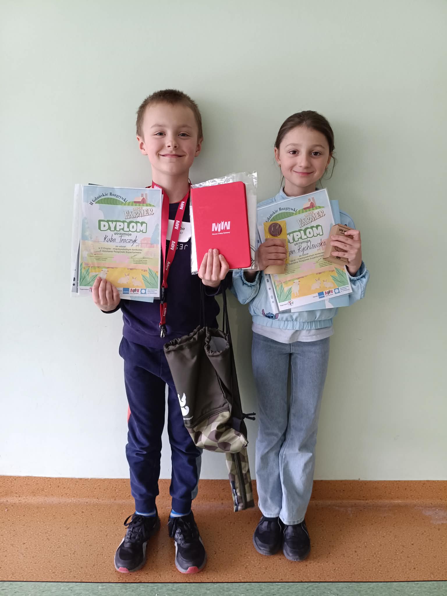 Dwoje uśmiechniętych dzieci stoi przy jasnej ścianie, trzymając dyplomy i nagrody. Chłopiec po lewej ma na szyi medal i trzyma także czerwoną teczkę oraz worek. Dziewczynka po prawej trzyma dyplom, medal i małe pudełko. Oboje wyglądają na dumne z osiągnięć.