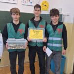 Trzech chłopaków w kamizelkach wolontariuszy stoi w sali lekcyjnej na tle tablicy szkolnej. Jeden z nich trzyma w rękach duże pudełko wypełnione monetami. Drugi ma w rękach napis Wrzuć grosika dla Smyka