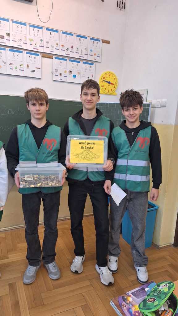 Trzech chłopaków w kamizelkach wolontariuszy stoi w sali lekcyjnej na tle tablicy szkolnej. Jeden z nich trzyma w rękach duże pudełko wypełnione monetami. Drugi ma w rękach napis Wrzuć grosika dla Smyka