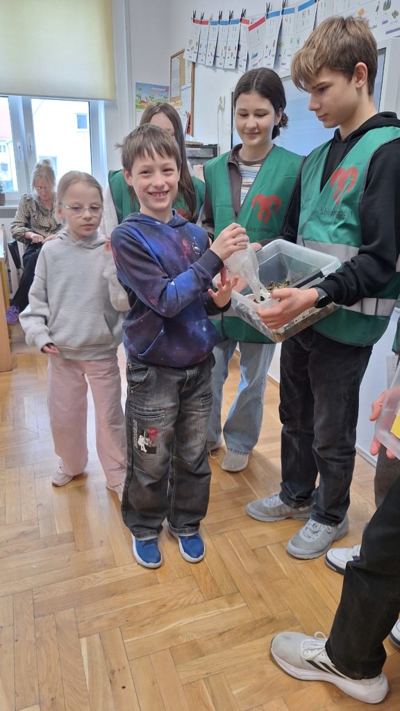 Grupa uczniów w klasie bierze udział w zbiórce pieniędzy – dzieci podchodzą kolejno i wrzucają monety do plastikowego pojemnika trzymanego przez starszych uczniów w kamizelkach wolontariusza
