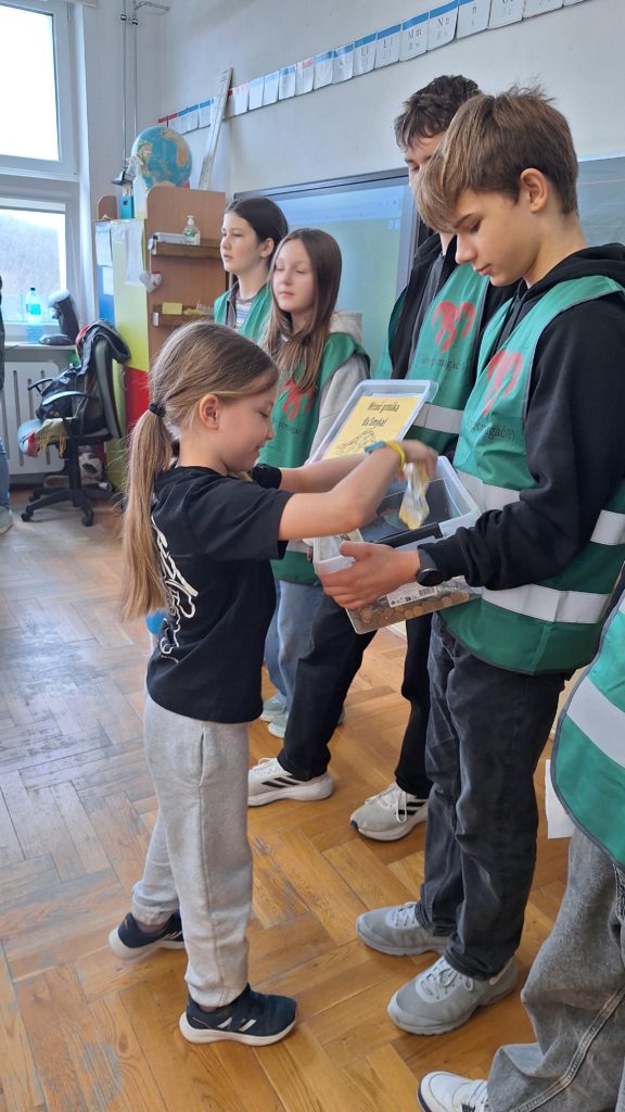 Grupa uczniów w klasie bierze udział w zbiórce pieniędzy – dzieci podchodzą kolejno i wrzucają monety do plastikowego pojemnika trzymanego przez starszych uczniów w zielonych kamizelkach wolontariusza.