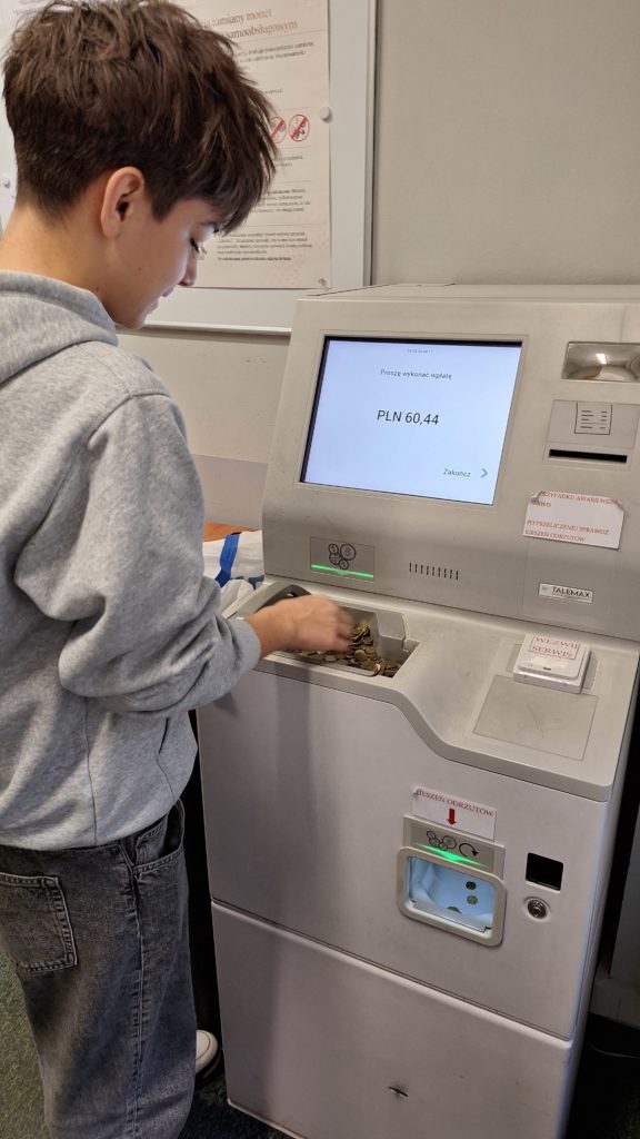 Chłopak stoi tyłem do robiącego zdjęcie. Wrzuca monety do maszyny liczącej. Na ekranie wyświetla się kwota przeliczonych pieniędzy.