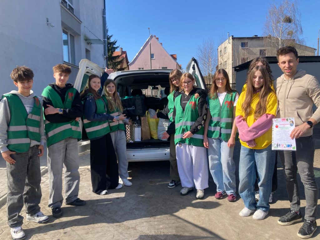 Grupa młodych wolontariuszy stoi przy otwartym bagażniku samochodu pełnym darów, uśmiechnięci na słonecznym podwórku między budynkami.