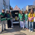 Grupa młodych wolontariuszy stoi przy otwartym bagażniku samochodu pełnym darów, uśmiechnięci na słonecznym podwórku między budynkami.