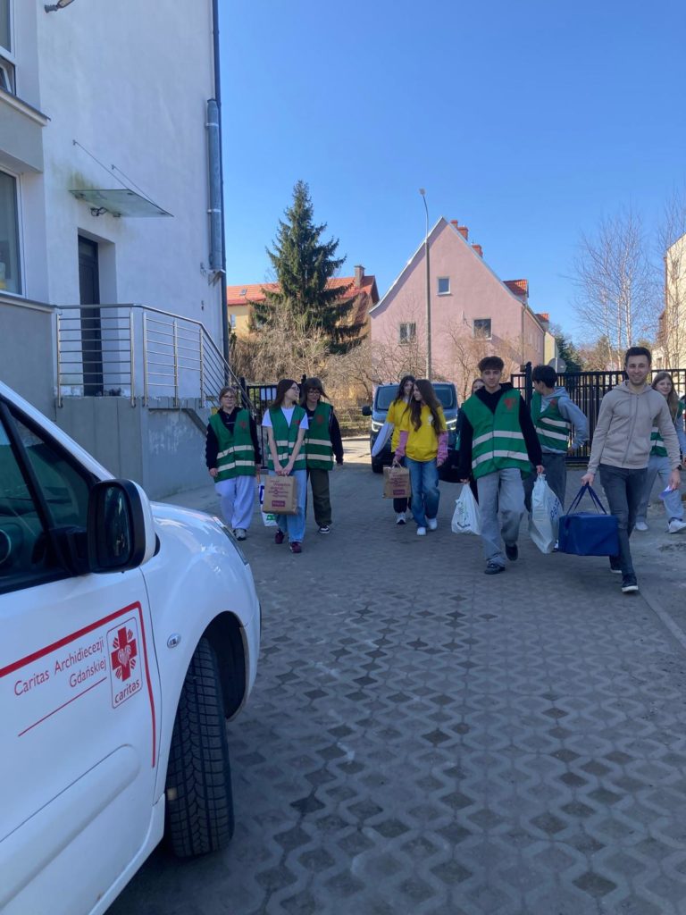 Grupa młodych wolontariuszy niesie torby z darami w stronę samochodu.