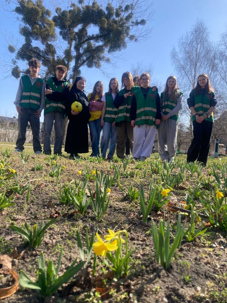 Młodzi wolontariusze stoją razem w ogrodzie z kwitnącymi żonkilami, pozując do zdjęcia w słoneczny dzień. Wśród nich jest siostra zakonna.