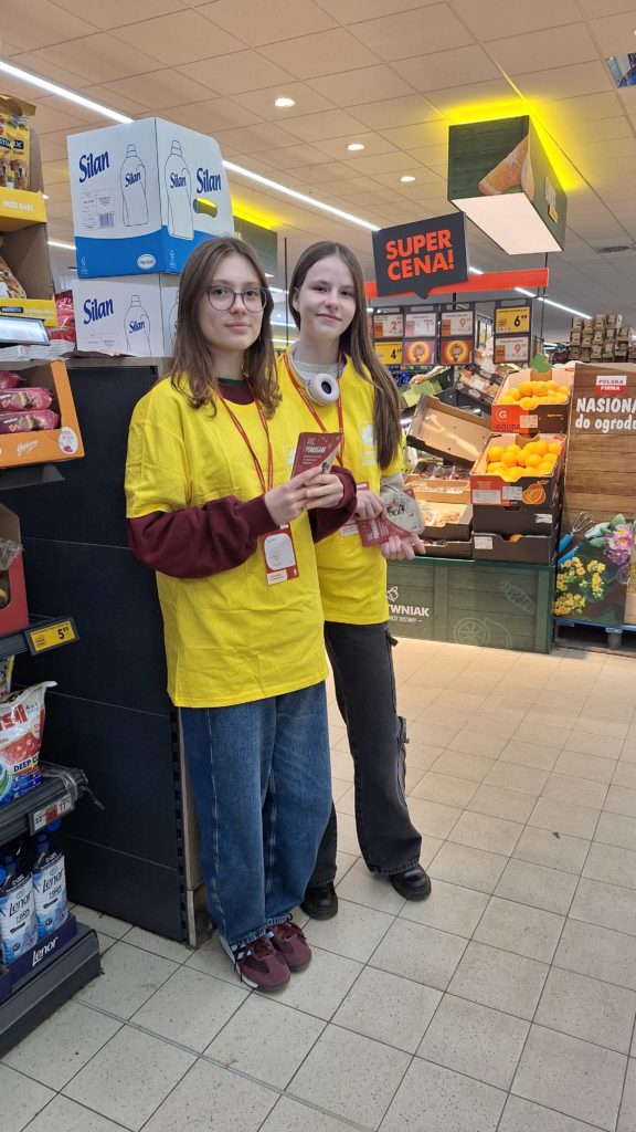 Dwie dziewczyny w koszulkach wolontariusza stoją na terenie sklepu biedronka i zachęcają klientów do udziału w zbiórce żywności. Trzymają w dłoniach ulotki. Są uśmiechnięte. W tle owoce i półki z produktami.