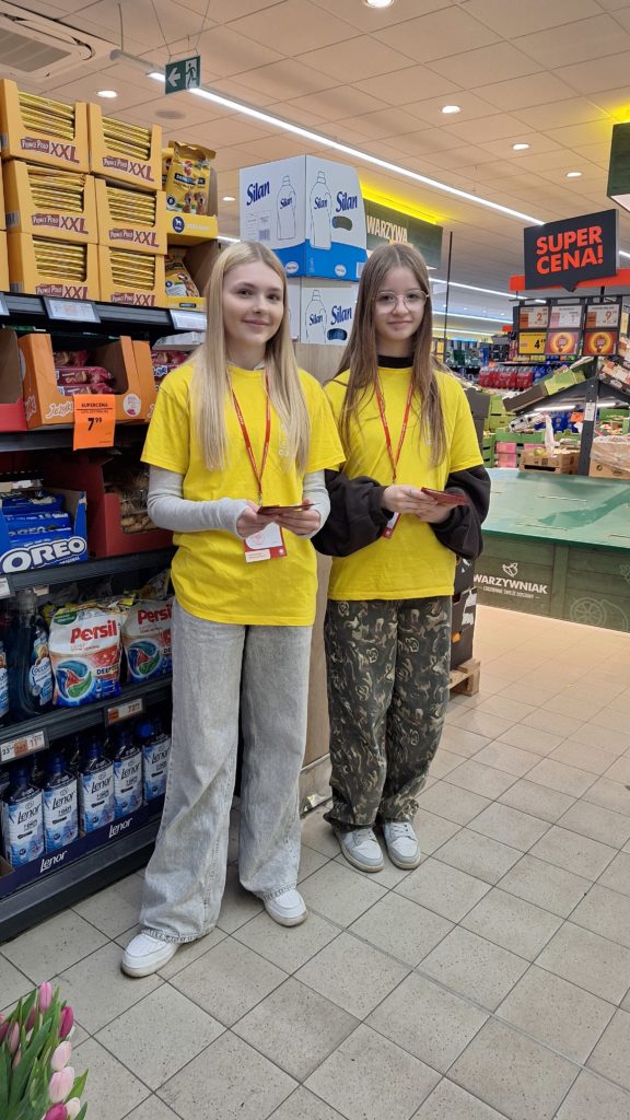 Dwie dziewczyny w koszulkach wolontariusza stoją na terenie sklepu biedronka i zachęcają klientów do udziału w zbiórce żywności. Trzymają w dłoniach ulotki. Są uśmiechnięte. W tle owoce i półki z produktami.