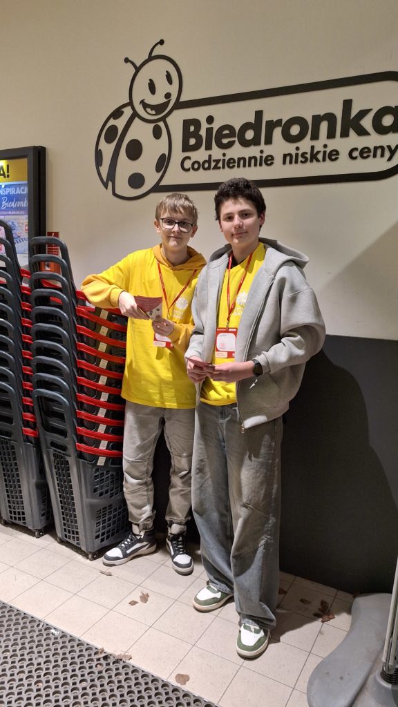 Dwóch chłopaków w koszulkach wolontariuszy stoi przy wejściu sklepu obok koszyków zakupowych i rozdaje ulotki.