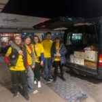 Pięć osób w koszulkach wolontariuszy stoi przy otwartym bagażniku dużego samochodu, którym znajdują się kartony. Jest wieczór.