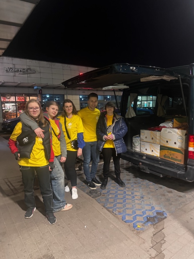 Pięć osób w koszulkach wolontariuszy stoi przy otwartym bagażniku dużego samochodu, którym znajdują się kartony. Jest wieczór.