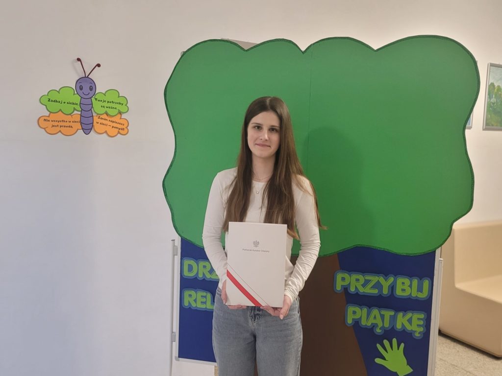 Małgosia stoi na tle dużego drzewa z papieru i prezentuje dyplom finalisty konkursu.