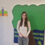 Małgosia stoi na tle dużego drzewa z papieru i prezentuje dyplom finalisty konkursu.