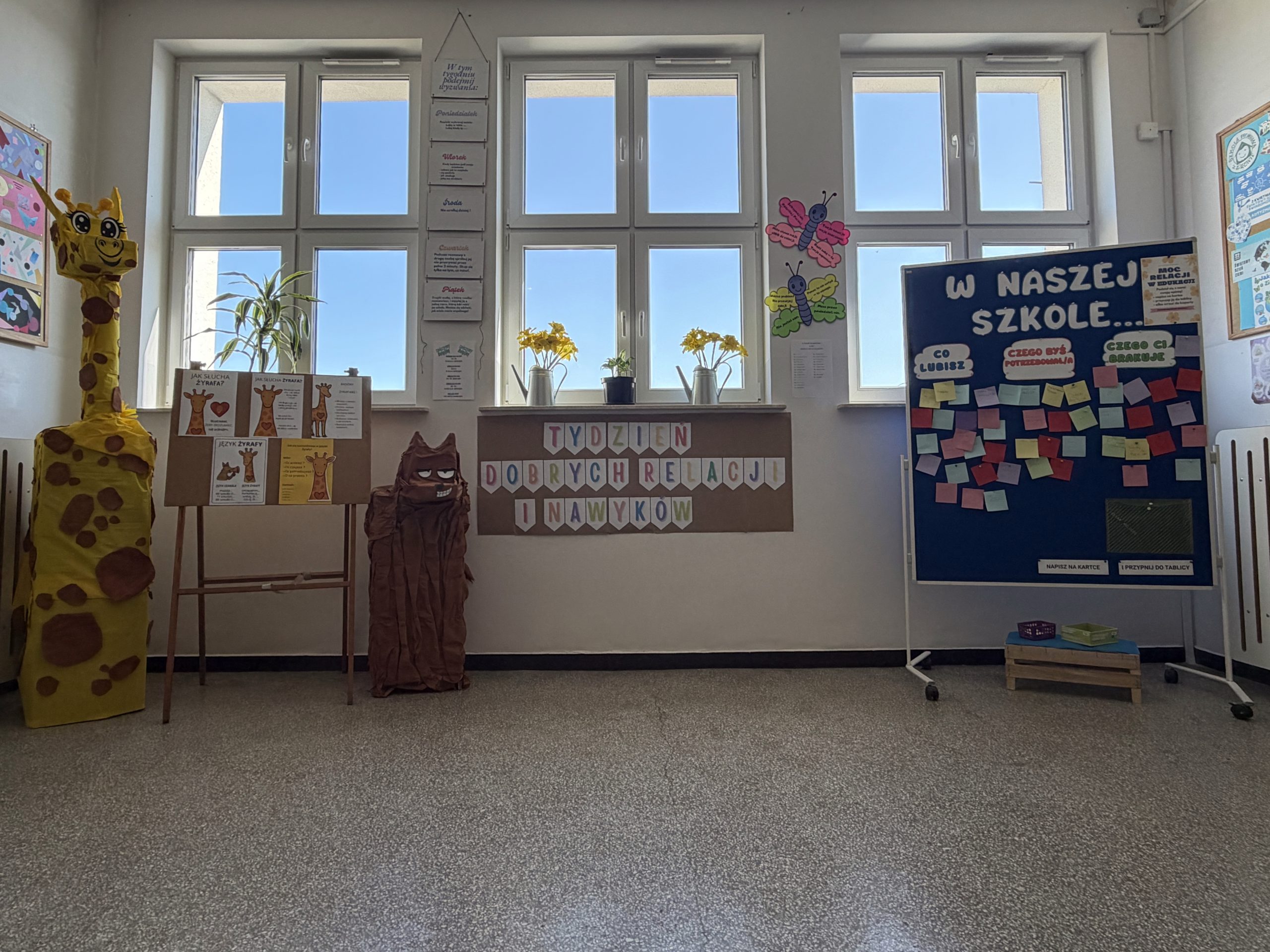 udekorowany korytarz szkolny. Na pierwszym planie po lewej stronie stoi duża żyrafa wykonana z papieru i kartonu, a obok niej tablica informacyjna na sztaludze. Na środku, na ścianie pod oknami, wisi baner z kolorowymi literami tworzącymi napis: „TYDZIEŃ DOBRYCH RELACJI I NAWYKÓW”. Po prawej stronie widać niebieską tablicę na kółkach z pytaniami w chmurkach: „CO LUBISZ”, „CZEGO BYŚ POTRZEBOWAŁ/A”, „CZEGO CI BRAKUJE”. Na tablicy są przypięte liczne kolorowe karteczki samoprzylepne.