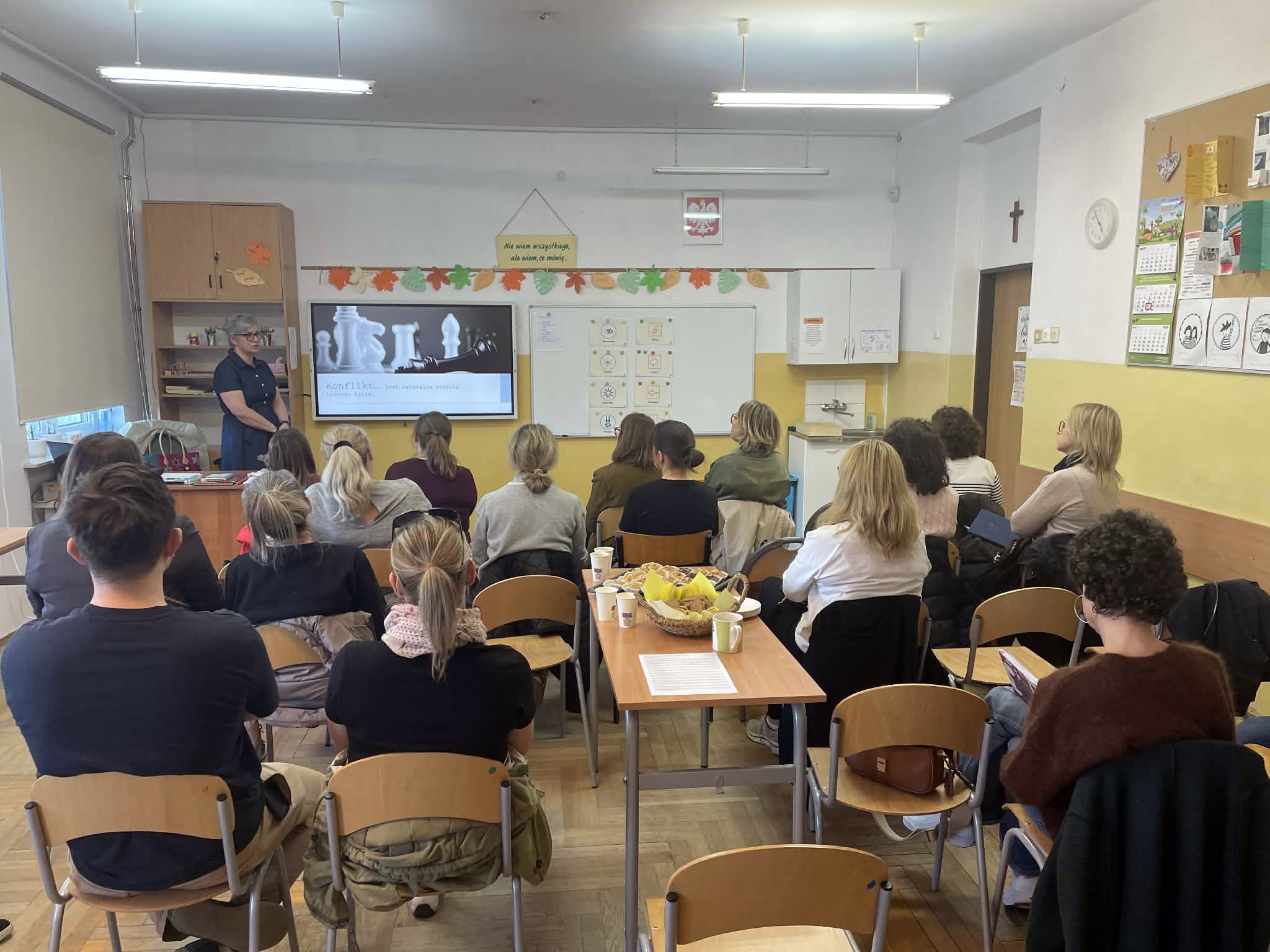Uczestnicy szkolenia siedzą w ławkach i patrzą na prezenterkę oraz ekran telewizora, na którym wyświetlany jest slajd z napisem „Konflikt... to naturalna część życia”. Na stole pośrodku stoją przekąski. Na tablicy wiszą karty z piktogramami.