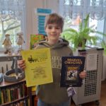 W centralnej części fotografii na tle regału bibliotecznego z książkami stoi uśmiechnięty  chłopiec  trzymający dyplom za zajęcie pierwszego miejsca i książkę, a z kieszeni jego bluzy wystaje czekolada. 