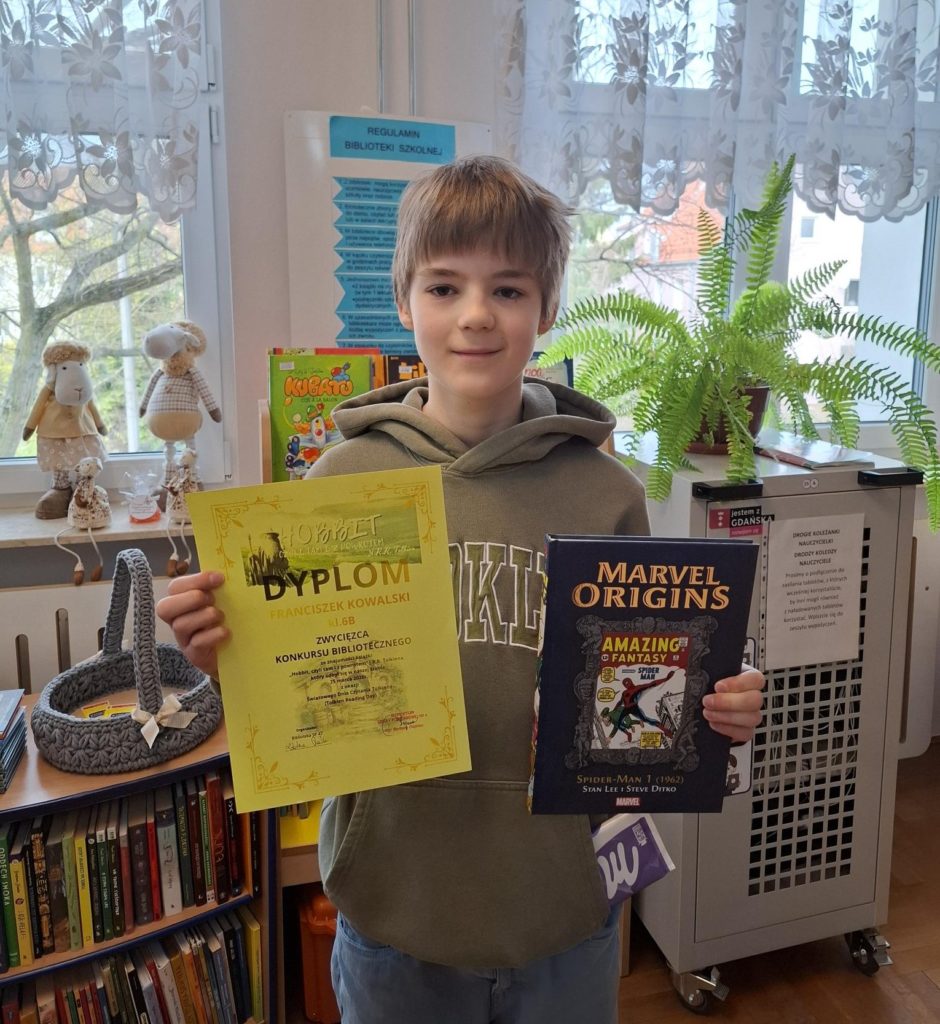 W centralnej części fotografii na tle regału bibliotecznego z książkami stoi uśmiechnięty  chłopiec  trzymający dyplom za zajęcie pierwszego miejsca i książkę, a z kieszeni jego bluzy wystaje czekolada. 