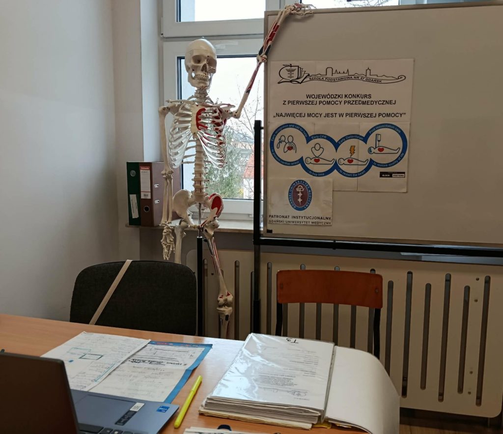 Na pierwszym planie biurko z otwartym laptopem i rozłożonymi dokumentami. W tle tablica z plakatem Wojewódzkiego Konkursu z Pierwszej Pomocy, ilustracją łańcucha przeżycia oraz logiem GUMed. Obok stoi model ludzkiego szkieletu.