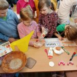 Grupa dzieci w klasie przeprowadza prosty eksperyment przy stole – jedno dziecko wlewa płyn do małego pojemnika, inne obserwuje i zapisuje wyniki w ćwiczeniach, a na blacie leżą przybory szkolne i materiały.