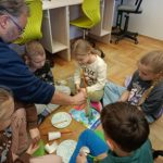 W kole na podłodze siedzi grupa dzieci i przygląda się wieży ze słomek, którą trzyma męż-czyzna. W koło słomki, kubeczki, talerzyki i kartki.